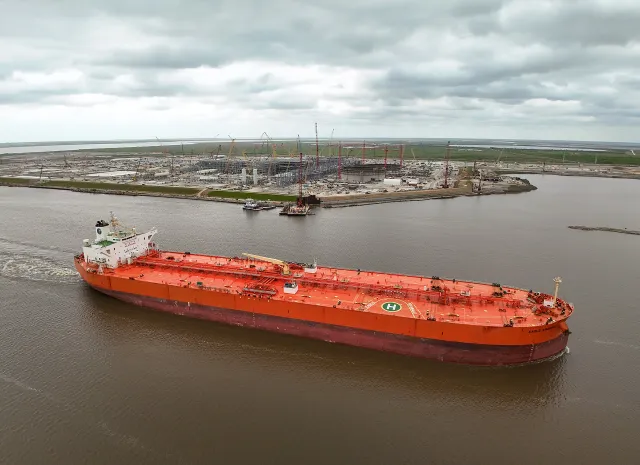 A red crude oil tanker sailing along a channel past an industrial site in Port Arthur, Texas