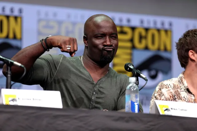 Mike Colter flexing his arm playfully while seated at a microphone on a San Diego Comic-Con panel